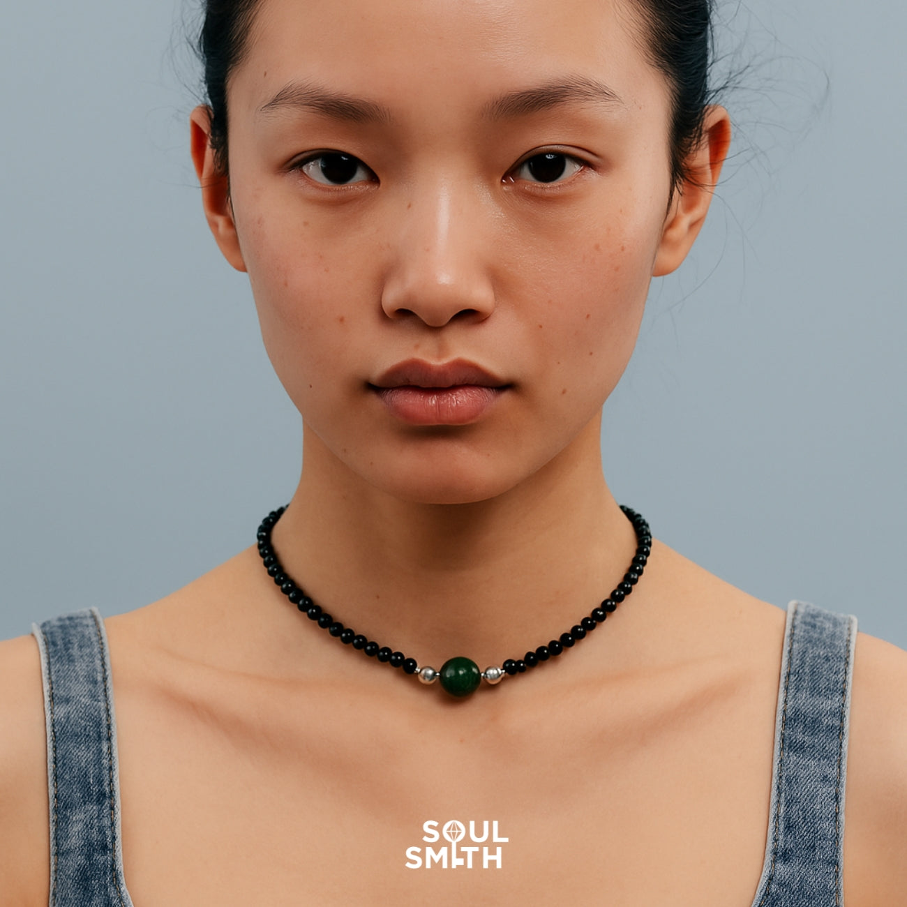 Close-up of a model wearing a SoulSmith choker necklace from the SoulLinks Collection, featuring small black beads, silver accents, and a single large green bead at the center, styled against a muted blue background with denim straps.