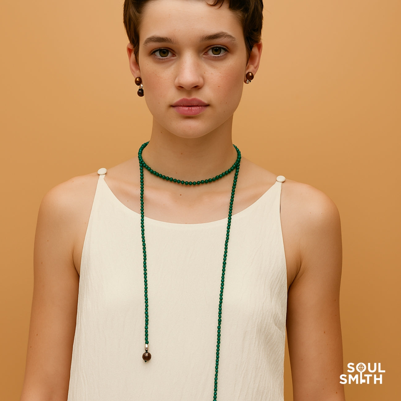 SoulSmith SoulLinks Collection — model wearing a green beaded rope necklace with tiger’s eye gemstone ends, styled around the neck and draped down over a white sleeveless top, against a soft peach background.