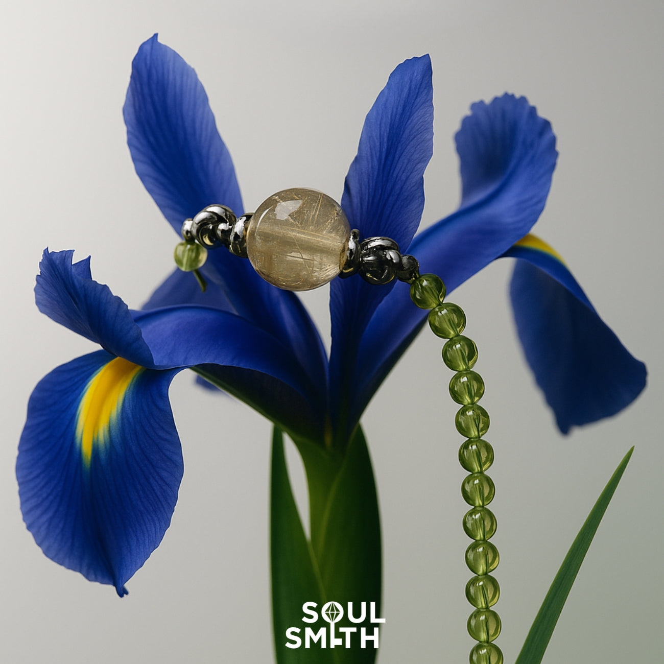 Golden Rutilated Quartz charm with golden threads, set between dark silver beads and green round peridot beads, elegantly draped over a vivid blue iris flower. SoulSmith branding below.