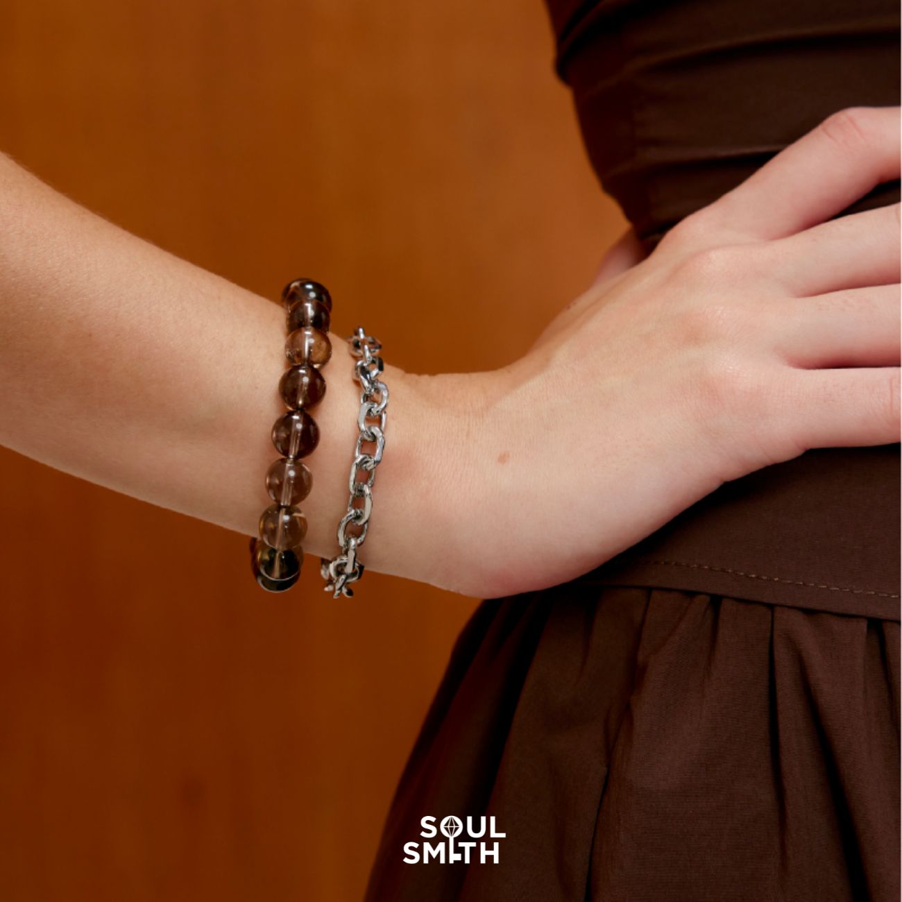 Close-up of a model wearing a Smoky Quartz crystal bracelet paired with a chunky silver chain bracelet, styled with a brown pleated dress