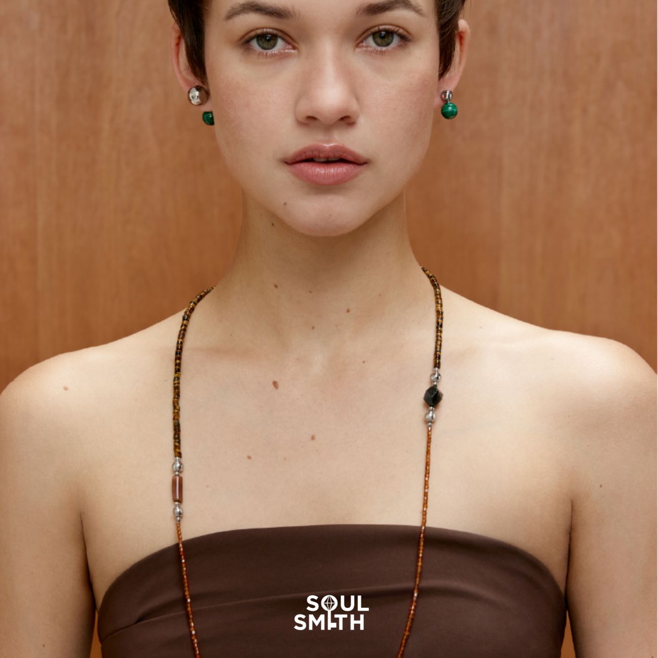 Woman wearing a long, tiger eye beaded rope necklace with black and tiger eye crystals, styled over a strapless brown top. The necklace features varied textures and colors, suggesting grounding energy. She pairs it with matching green gemstone earrings. Background is a soft wooden panel,
