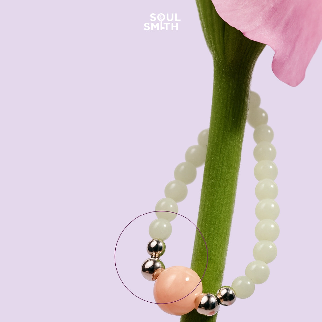 Close-up of a Soulsmith necklace with a soft pink Morganite centerpiece, surrounded by shiny silver beads and creamy round stones, draped elegantly around a green flower stem on a pastel lavender background.
