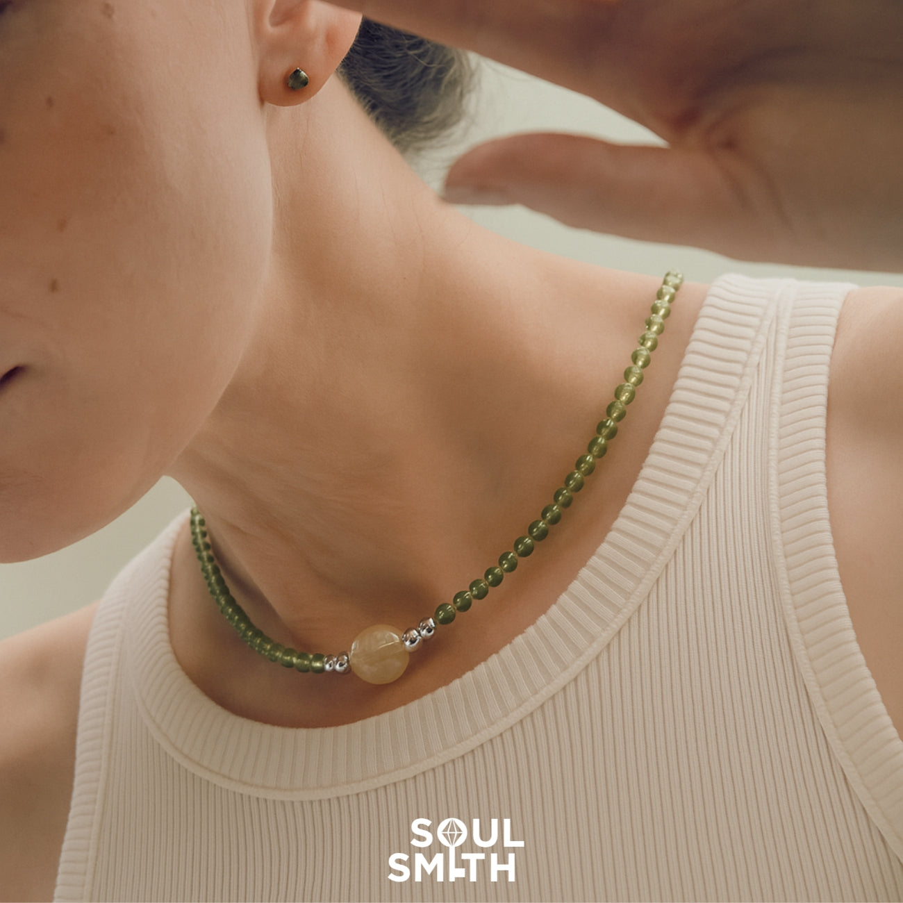Close-up of a woman’s neck wearing a SoulSmith necklace made with golden rutilated quartz as the central charm, accented by polished green peridot beads and silver details, styled with a ribbed cream tank top.