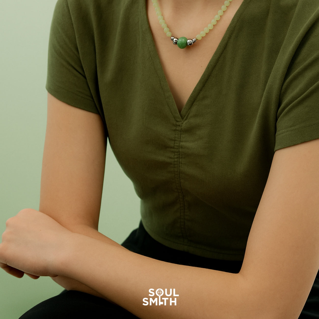Woman wearing a green V-neck top styled with a round Berylite gemstone charm necklace, accented with silver beads and pastel green beading, captured in a minimalist and serene setting for Soul Smith.