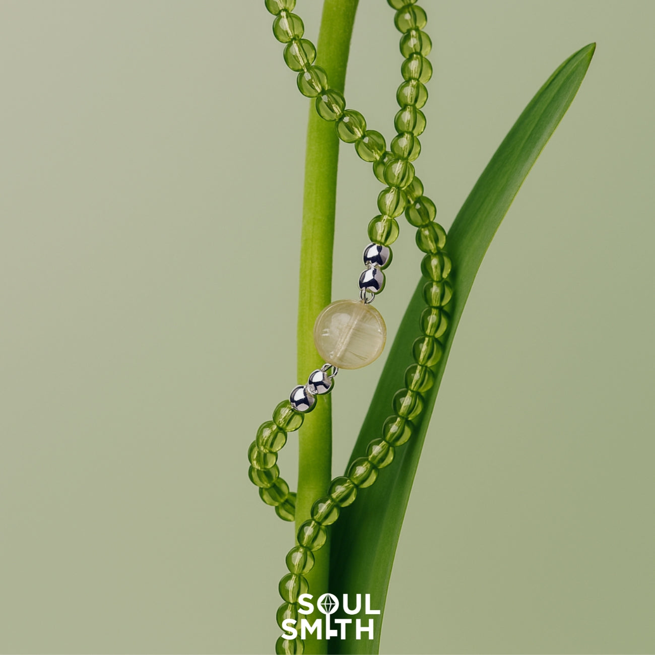 SoulSmith necklace featuring a golden rutilated quartz centerpiece with silver accents, strung on translucent green peridot beads, elegantly wrapped around a fresh green plant stem on a pale green background.