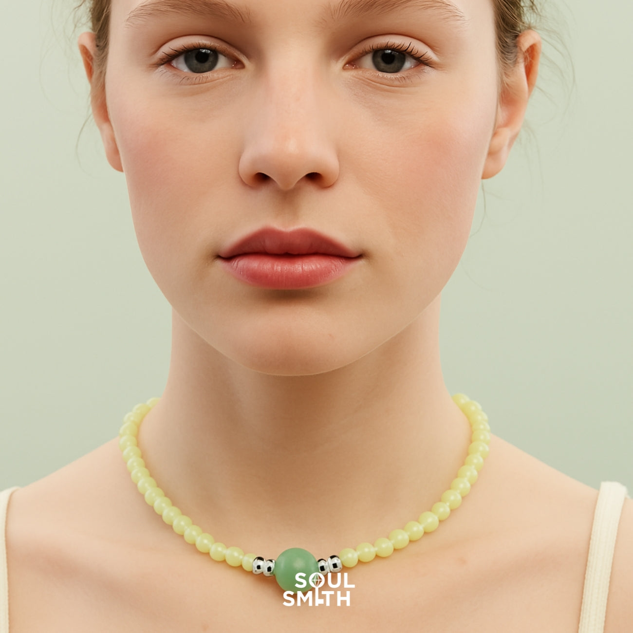 Woman wearing a soft yellow beaded necklace with a centered Berylite gemstone charm and metallic accents, styled against a pale green backdrop for Soul Smith.