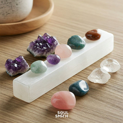 Tumbled chakra crystals arranged on a selenite charging bar, featuring amethyst clusters, rose quartz, green aventurine, clear quartz, and more—displayed on a wooden surface with the Soulsmith logo.
