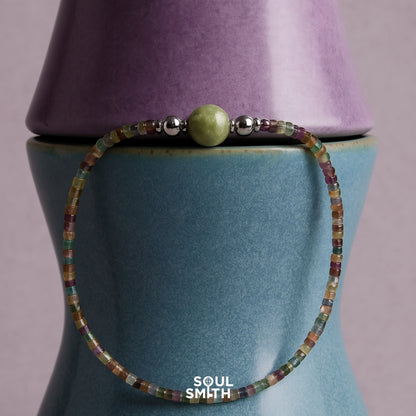Minimalist bracelet with a Berylite center bead and multicolored square-cut gemstone strand, styled on stacked ceramic vases.

