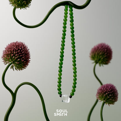 Jade beaded necklace with a square-cut blue needle clear quartz centerpiece and black accent beads, artfully suspended among sculptural allium flowers — a statement piece from the Soulsmith SoulLinks collection.
