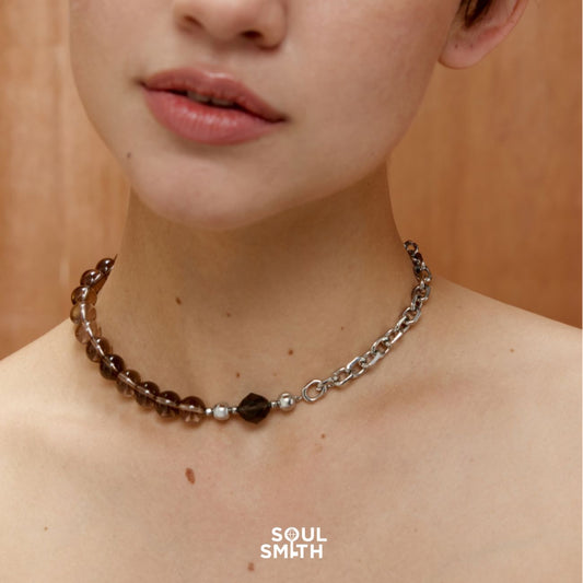 Close-up of a woman wearing a choker-style necklace composed of round smoky quartz beads on one half and a silver chain on the other, with a faceted black Apache Tears Obsidian Charm. 
