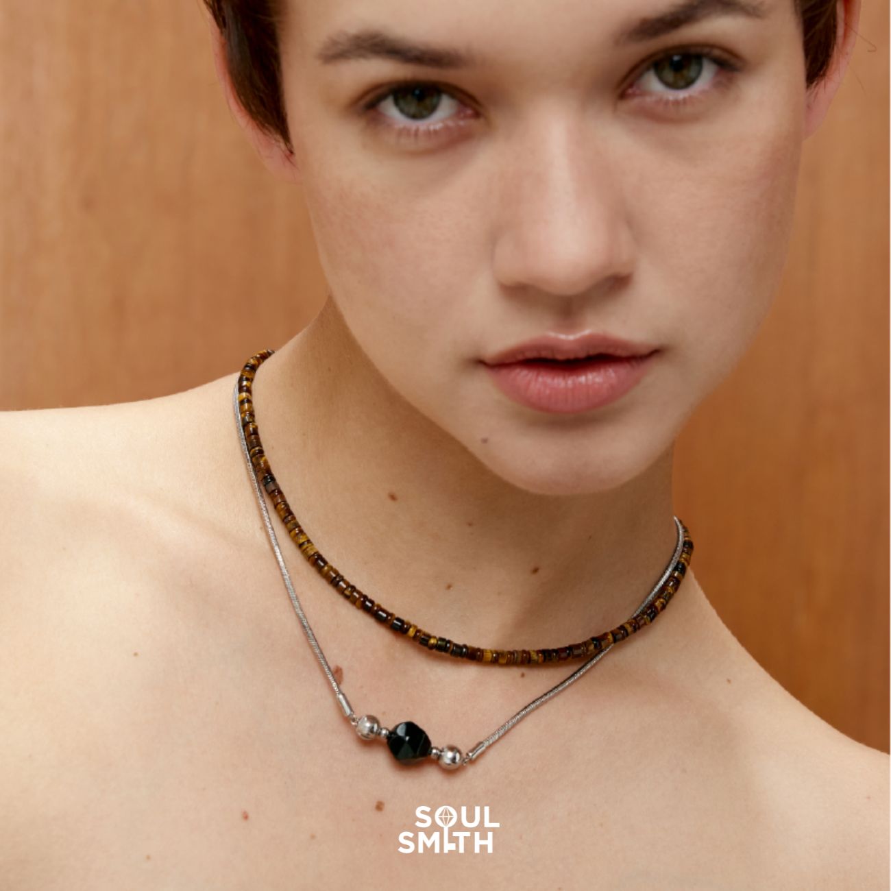 Close-up of a woman wearing a layered necklace set: one choker made of small, earthy tiger eye beads, and another featuring a Apache Tears Obsidian Charm on a silver chain. Her expression is serene, with a wooden background 