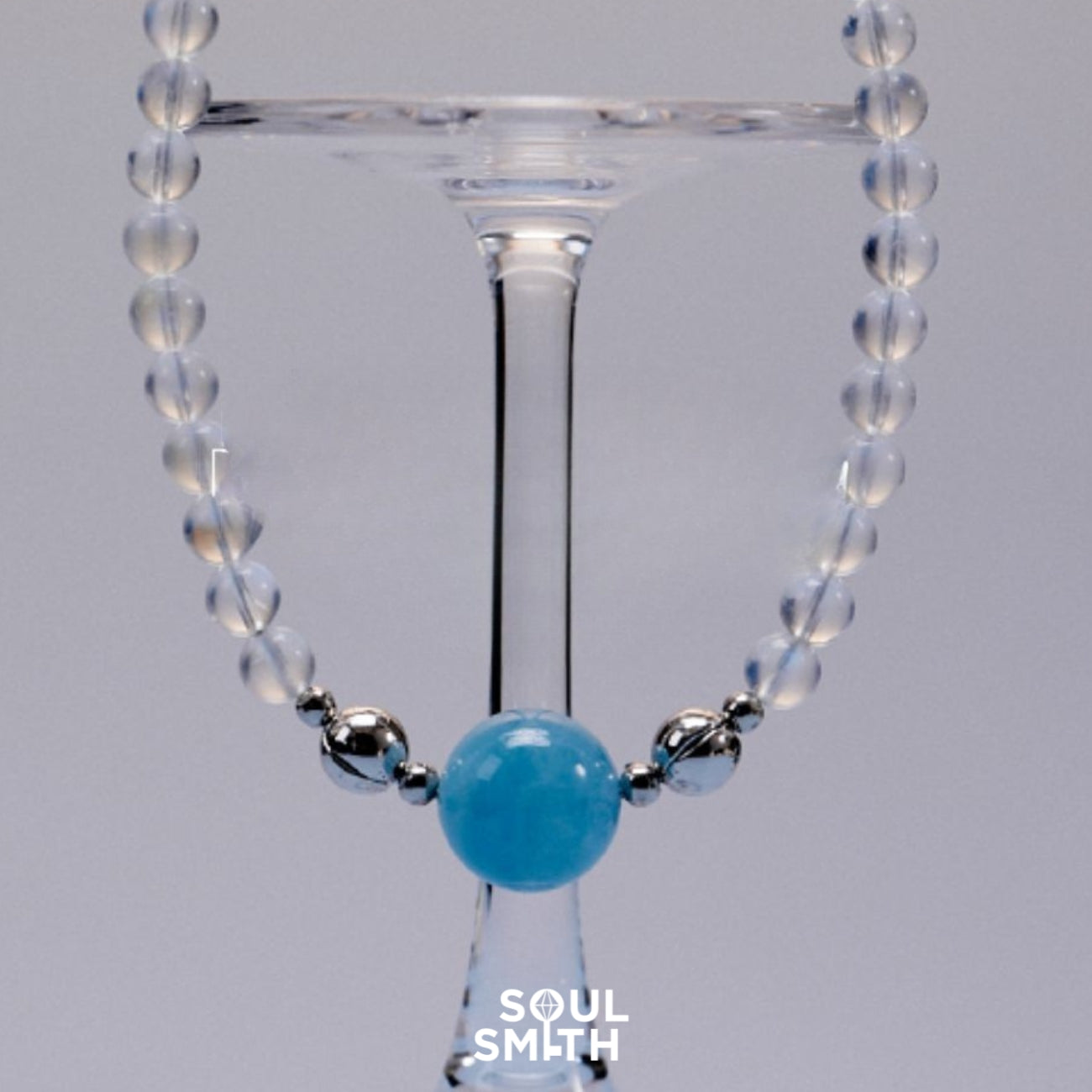 Aquamarine charm necklace with translucent moonstone beads and silver accents, elegantly draped over a wine glass, reflecting a cool and serene aesthetic.
