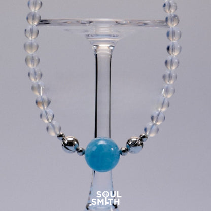Aquamarine charm necklace with translucent moonstone beads and silver accents, elegantly draped over a wine glass, reflecting a cool and serene aesthetic.
