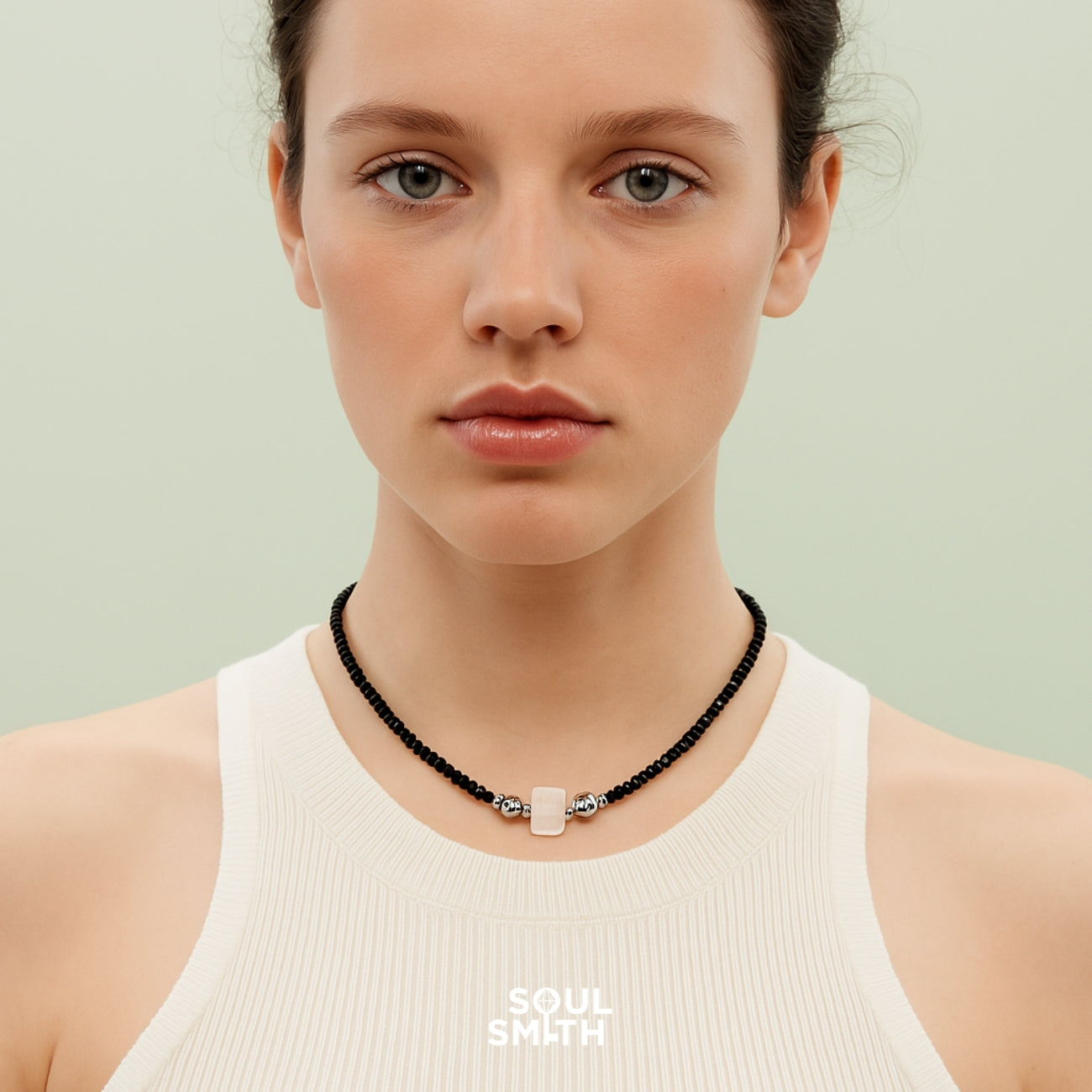Model wearing a minimalist black onyx beaded necklace featuring a rectangular blue needle crystal charm with silver accents, styled with a white ribbed tank top against a soft mint green background — part of the Soulsmith SoulLinks collection.