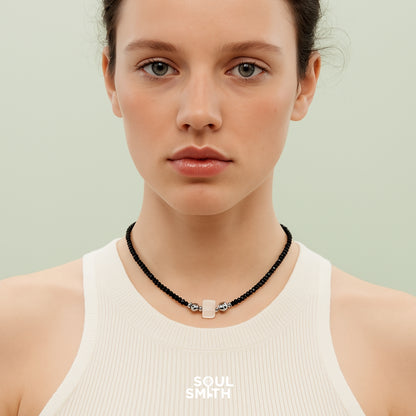 Model wearing a minimalist black onyx beaded necklace featuring a rectangular blue needle crystal charm with silver accents, styled with a white ribbed tank top against a soft mint green background — part of the Soulsmith SoulLinks collection.