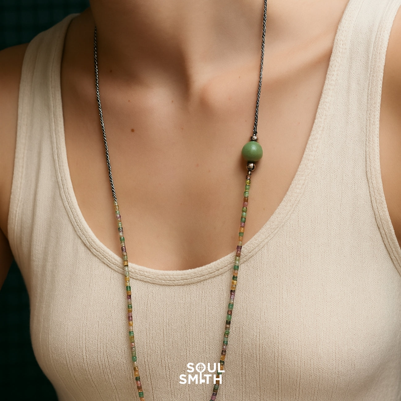 Long handcrafted necklace featuring a green Berylite charm paired with multicolored square-cut beads and a twisted rope chain, worn over a beige ribbed tank.
