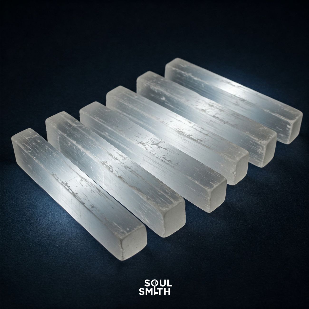 Six polished selenite crystal sticks aligned on a dark surface, glowing softly with white light. Ideal for spiritual cleansing, chakra work, and crystal recharging – Soulsmith branding included.
