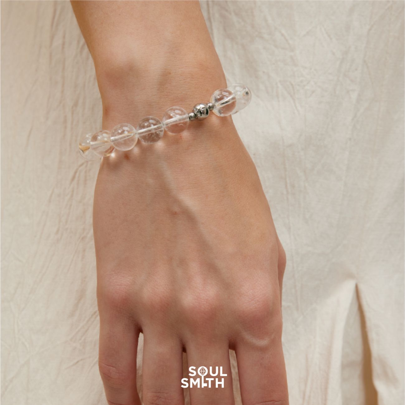 Close-up of a hand wearing a Soulsmith Clear Quartz crystal bracelet with large round beads and a magnetic clasp, styled against a soft beige dress for a clean, spiritual look.
