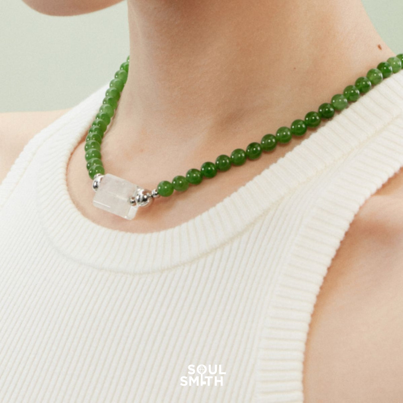 Close-up of a model wearing a Soulsmith necklace featuring a blue needle crystal rectangular charm, flanked by silver beads and strung on vibrant green round beads, styled with a white ribbed tank top.
