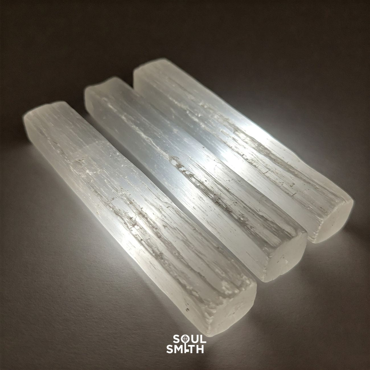 Three polished selenite crystal sticks glowing under light on a neutral background, used for energy cleansing and recharging other crystals – Soulsmith branding visible.
