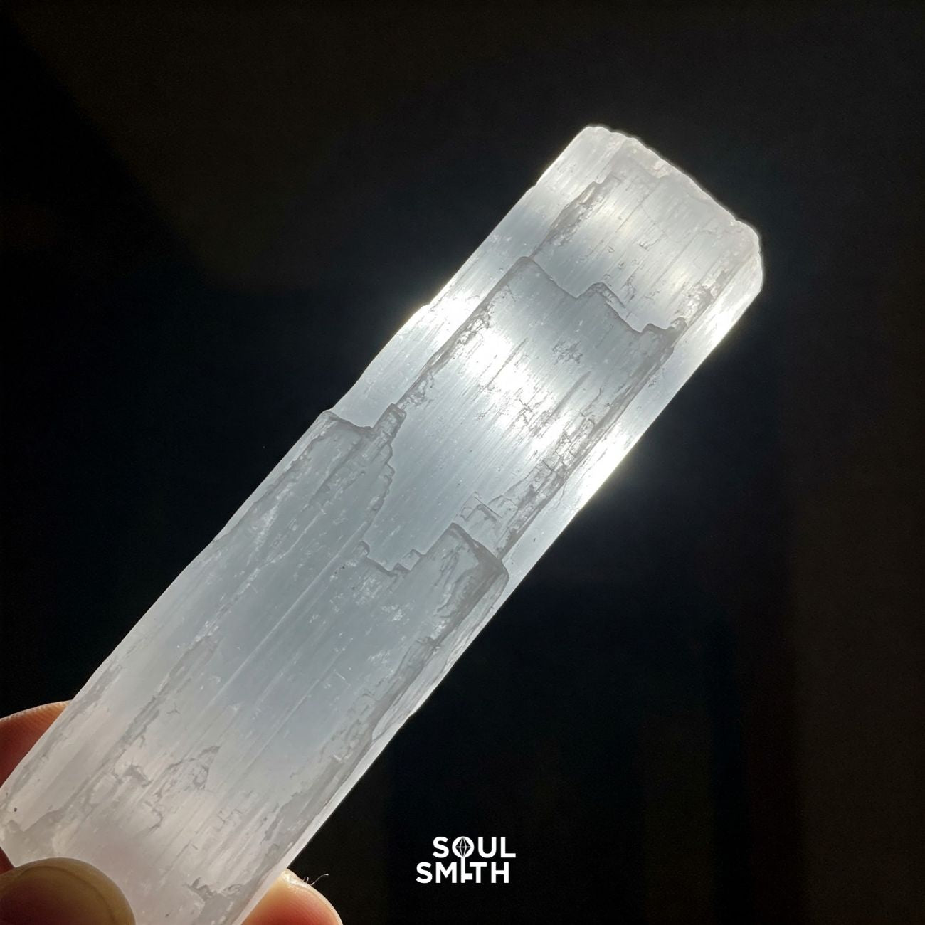 Hand holding a raw selenite crystal stick glowing under light, symbolizing energy cleansing and spiritual purification – Soulsmith branding visible.
