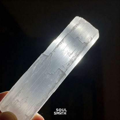 Hand holding a raw selenite crystal stick glowing under light, symbolizing energy cleansing and spiritual purification – Soulsmith branding visible.
