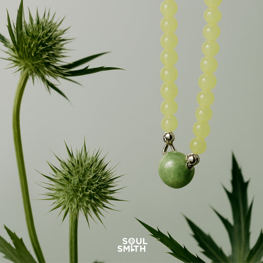 Green Berylite pendant paired with soft yellow round beads, suspended delicately against spiky green thistle plants on a minimal pale backdrop, styled for Soul Smith.
