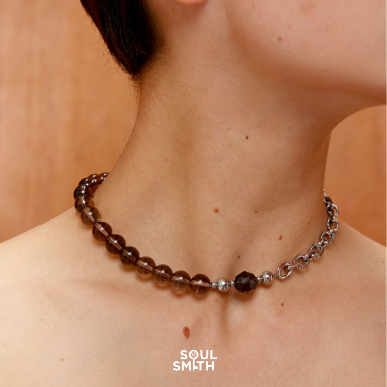 Close-up of a woman’s neck wearing a Smoky Quartz crystal necklace featuring round translucent brown beads on one side, an Apache Tears Obsidian Charm at the center, and a chunky silver chain on the other side. The piece balances raw earthiness with bold, edgy style. The background is a warm wood tone, and the Soul Smith logo is visible at the bottom.
