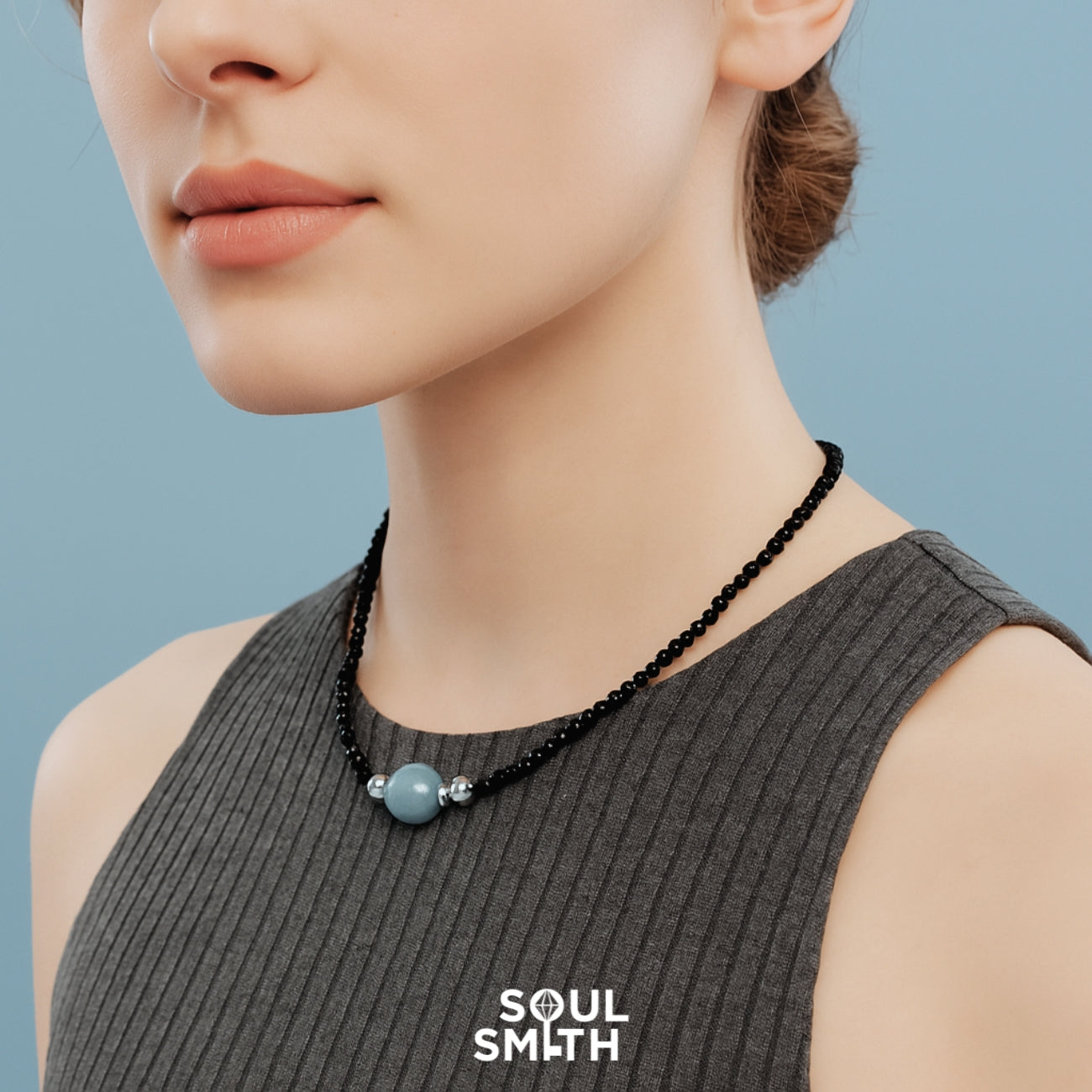 Woman wearing a ribbed charcoal-grey sleeveless top and a delicate black beaded necklace centered with a soft blue aquamarine charm and silver accents — a SoulLinks piece by Soulsmith.
