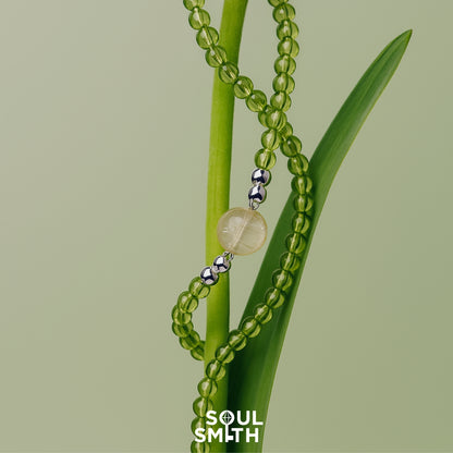SoulSmith necklace featuring a golden rutilated quartz centerpiece with silver accents, strung on translucent green peridot beads, elegantly wrapped around a fresh green plant stem on a pale green background.
