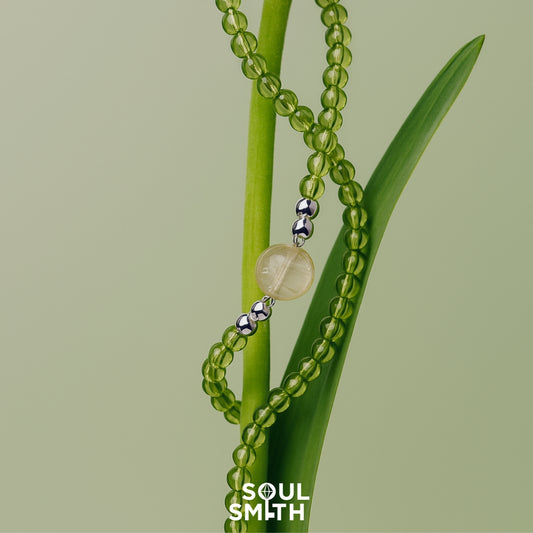 SoulSmith necklace featuring a golden rutilated quartz centerpiece with silver accents, strung on translucent green peridot beads, elegantly wrapped around a fresh green plant stem on a pale green background.
