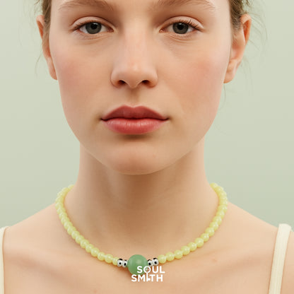 Woman wearing a soft yellow beaded necklace with a centered Berylite gemstone charm and metallic accents, styled against a pale green backdrop for Soul Smith.
