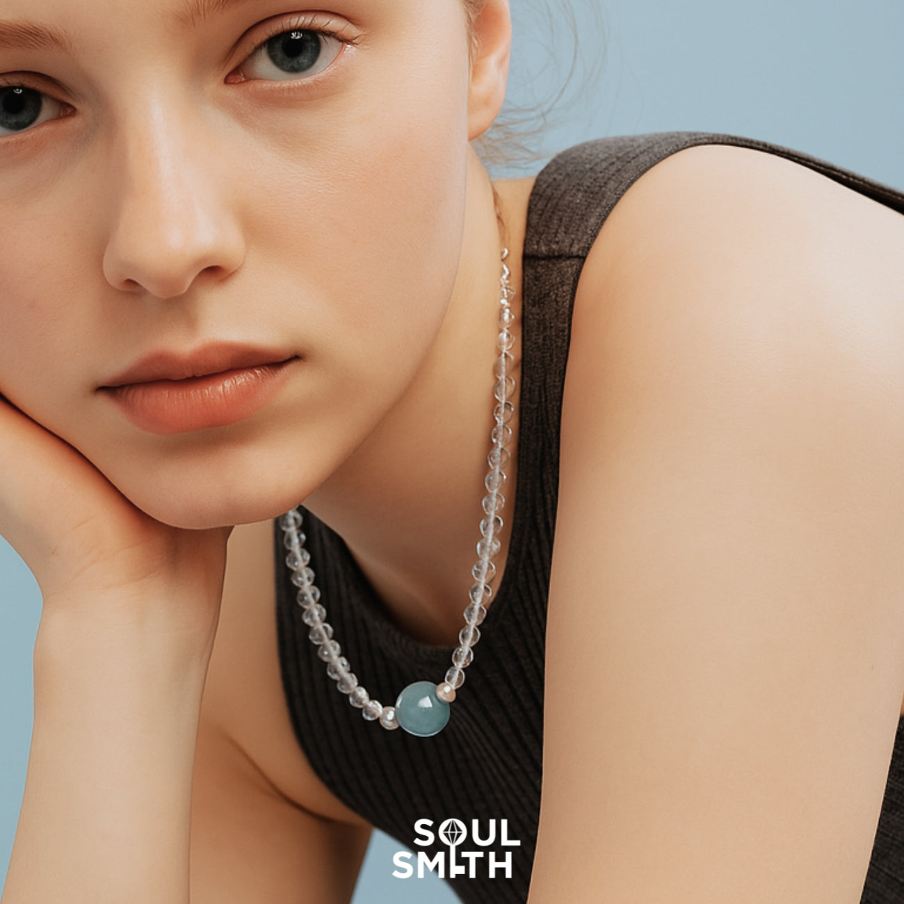 Young woman resting her face on her hand, wearing a dark sleeveless ribbed top and a translucent crystal beaded necklace with a central aquamarine charm — a calming handcrafted SoulLinks piece from Soulsmith.
