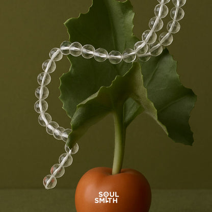 Soulsmith Clear Quartz crystal bead bracelet with polished 8mm transparent stones draped over a green leaf and terracotta base, symbolizing purity and crown chakra clarity.
