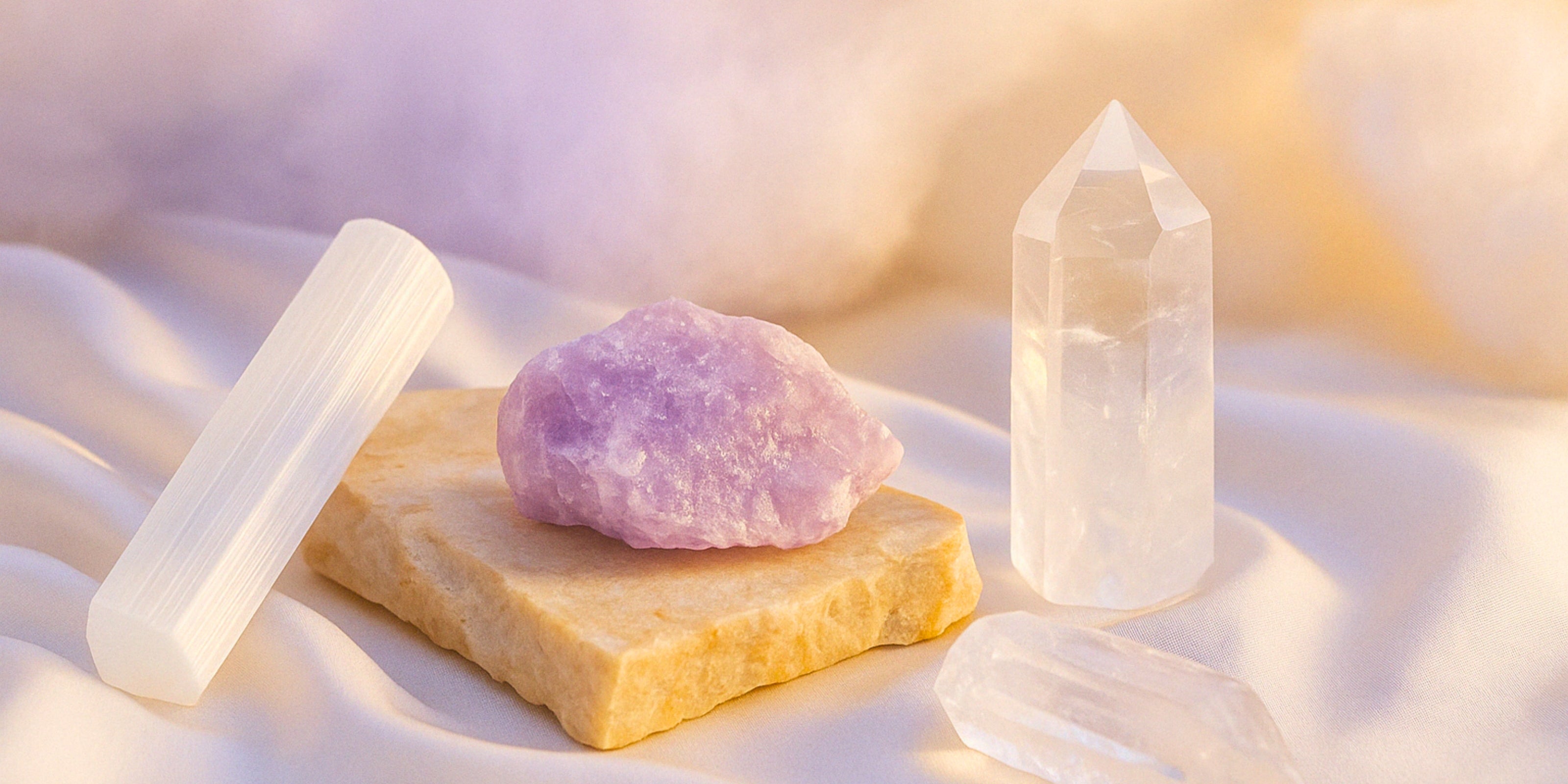 A dreamy trio of crystals: selenite wand (Crown Chakra), raw amethyst chunk (Third Eye Chakra), and a polished clear quartz tower (Crown Chakra), arranged on satin fabric with warm, glowing light for an ethereal healing vibe.
