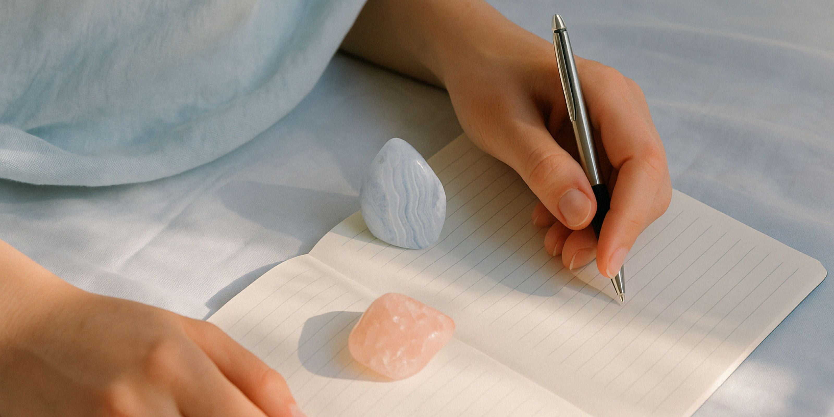 Hand writing in journal beside blue lace agate and rose quartz crystals on soft blue fabric