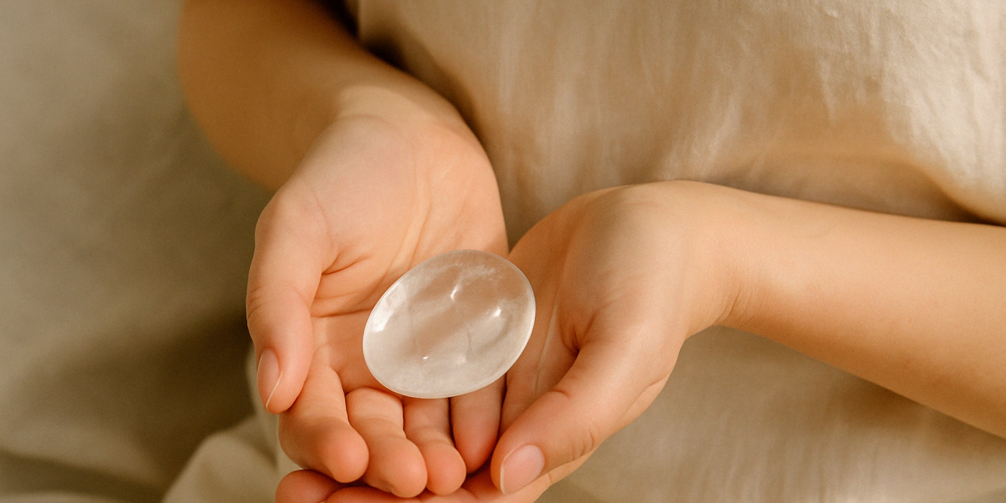 Hands gently holding a polished clear quartz palm stone against neutral fabric